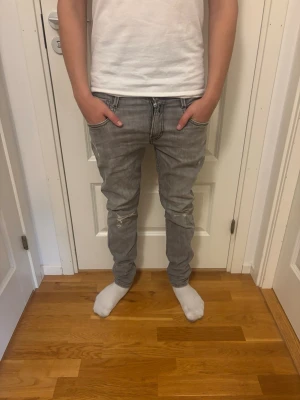 Gråa slim fit jeans med slitningar - Snygga gråa slim fit jeans med slitningar vid knäna och coola detaljer på bakfickorna. Jeansen har klassisk femficksmodell och dragkedjegylf. Perfekta för en avslappnad och trendig look.
