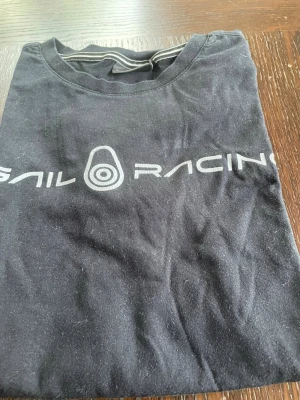 Svart Sail Racing t-shirt - Snygg svart t-shirt från Sail Racing med tryckt logga i vitt framtill. Klassisk rund hals och korta ärmar. Perfekt till jeans eller shorts för en avslappnad look.