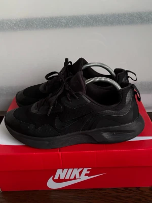 Svarta Nike sneakers i mesh och skinn - Snygga helsvarta Nike sneakers med lågt skaft. Ovandelen är i en mix av mesh och syntetiskt skinn för både stil och komfort. Klassisk Nike-logga på sidan och tungan. Perfekta för dig som gillar en clean och sportig look.