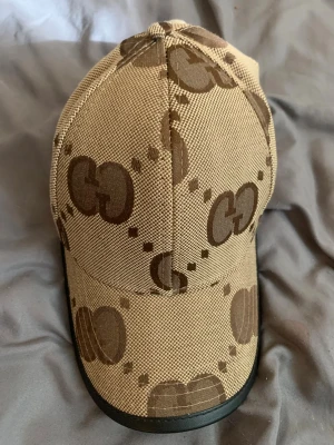 Beige keps med GG-mönster - Snygg beige keps med brunt GG-mönster över hela och svart kant på skärmen. Kepsen har klassisk form och är tillverkad i ett tåligt tygmaterial. Perfekt för dig som vill ha en trendig accessoar med streetkänsla.