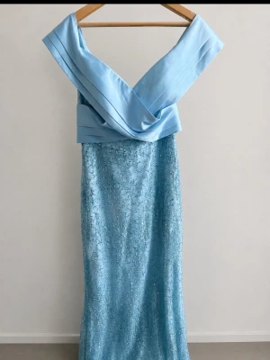 Ljusblå långklänning med paljetter oufshoulder - Elegant ljusblå långklänning med glittriga paljetter och bred omlottdetalj över axlarna. Klänningen har en figurnära passform och är perfekt för speciella tillfällen. Materialet ser ut att vara polyester eller liknande med skimrande yta. Pris kan diskuteras💙 