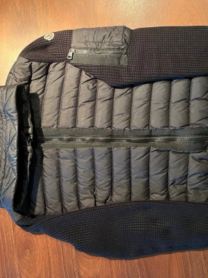 Äkta Moncler cardigan -   Moncler Cardigan Tricot i svart, herrmodell storlek L.  Padded/dunfodrad front och huva med stickade ärmar. Moncler-logga på ärmen.  Modell: F20919B50800 A9340 Storlek: L Färg: Svart  Fint begagnat skick. Mindre nötning/slitage på insidan vid dragkedjan, se bild. Syns inte när jackan är stängd. I övrigt fint skick.  Original dustbag och tags finns kvar. NFC-taggen fungerar. Tar bud
