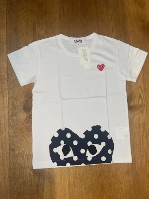 Comme des Garçons PLAY t-shirt - Vit t-shirt från Comme des Garçons PLAY med det ikoniska röda hjärtat broderat på bröstet och ett stort svartvitt prickigt hjärtmotiv nedtill. Klassisk rund hals och korta ärmar. Perfekt för dig som gillar streetwear och vill sticka ut. Säljer nu denna Comme des Garçons T-shirt som blev för liten för mig KÖPT PÅ SELLPY