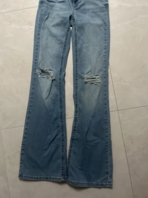 Ljusblå bootcut jeans med slitningar - Säljer ett par ljusblå bootcut jeans med slitna detaljer över knäna och snygg broderad ficka bak. Jeansen har klassisk femficksmodell, bälteshällor och är tillverkade i denim. Perfekta för dig som gillar en avslappnad och trendig stil.