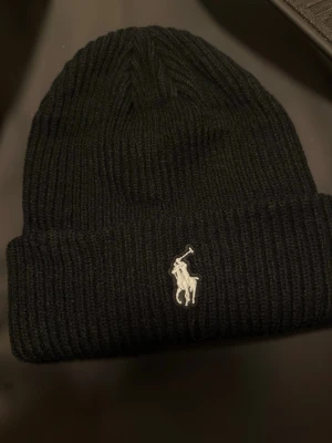 Svart  mössa Polo Ralph Lauren - Säljer en svart ribbstickad mössa från Polo Ralph Lauren med den klassiska broderade loggan framtill. Mössan har uppvikt kant och är tillverkad i mjukt material som håller dig varm under kalla dagar. Perfekt accessoar för en stilren look.