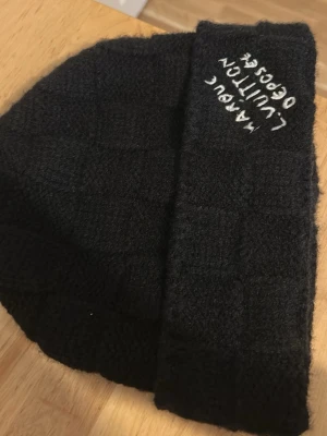 Svart stickad mössa med brodyr - Säljer en svart stickad mössa med rutmönster och broderad text på uppviket. Mössan har en klassisk form och är gjord i ett mjukt, värmande material som passar perfekt för kalla dagar. Enkel men ändå unik detalj med brodyren framtill.