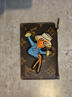 Louis Vuitton plånbok med patch - Cool brun plånbok från Louis Vuitton med klassiskt monogrammönster och en färgglad patch av en tecknad figur i blå kostym och vit hatt. Plånboken har guldfärgad dragkedja med LV-logga och är tillverkad i läder.