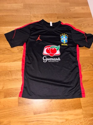 Svart Brasilien fotbollströja - Snygg svart fotbollströja med röda detaljer och Brasilien-emblem på bröstet. Tröjan har Guaraná Antarctica-logga framtill och gult BRASIL-tryck på ryggen. Tillverkad i ett lätt och ventilerande material, perfekt för träning eller match.