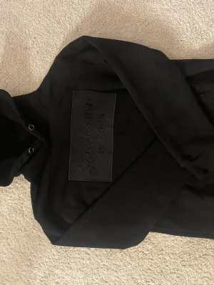 Svart hoodie från Sandhamn Sweden - Snygg svart hoodie från Sandhamn Sweden med stor broderad logga på bröstet. Hoodien har huva och är tillverkad i mjukt material som passar perfekt till en avslappnad stil.