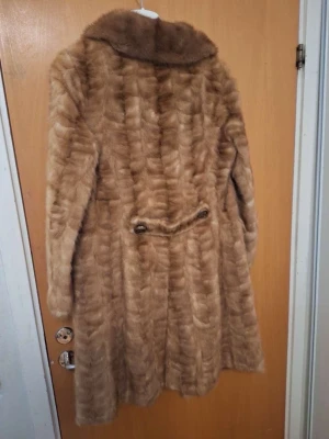 Lång brun fuskpäls kappa - Snygg lång kappa i brun fuskpäls med bred krage och knappar framtill. Kappan har två fickor och ett bälte bak för extra stil. Perfekt för kalla dagar när du vill vara både varm och trendig.