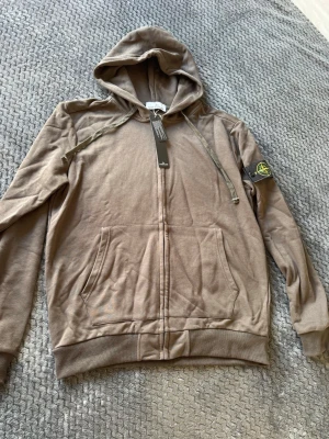 Brun hoodie med dragkedja och huva - Säljer en brun/grå hoodie med dragkedja och huva. Hoodien har fickor framtill, ribbade muddar och en patch med logga på vänster ärm. Tillverkad i mjukt bomullsmaterial och har justerbara snören i huvan. Perfekt för en avslappnad och stilren look.