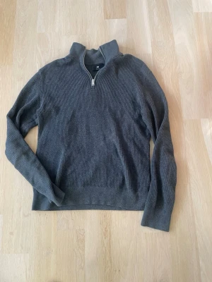 Mörkgrå stickad tröja med halv dragkedja - Mysig mörkgrå stickad tröja med hög krage och halv dragkedja framtill. Tröjan har lång ärm och ribbad struktur som ger en avslappnad men ändå stilren look. Perfekt för lager på lager under kyliga dagar.