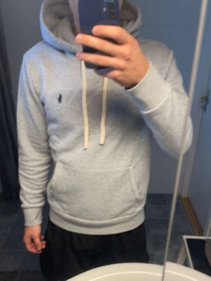 Ralph Lauren Hoodie - Snygg grå hoodie från Polo Ralph Lauren med klassisk huva och vita dragsnören. Tröjan har en känguruficka framtill och den ikoniska blå loggan broderad på bröstet. Tillverkad i mjukt material som känns skönt mot huden. Perfekt för en chill och stilren look. Taggarna och klistermärket har jag tagit av.