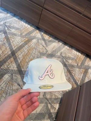 Grå Atlanta braves keps - Säljer en grå snapback-keps med Atlanta Braves-logga i vitt framtill och broderad 1995 World Series-detalj på sidan. Kepsen har platt skärm och små broderade hjärtan vid loggan. Klassisk streetwear-stil, perfekt för dig som gillar sportiga accessoarer.