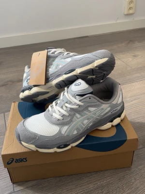 Gråa ASICS sneakers med mesh och skinn - Säljer ett par snygga ASICS sneakers i grått och vitt med klassisk logga på sidan. Skorna har en chunky sula i beige och svart, ovandel i mesh och detaljer i skinn. Perfekta för dig som gillar sportig och trendig stil. Snörning framtill för bästa passform.