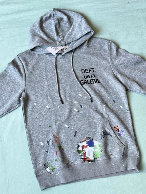 Grå hoodie Dept de la Galerie - HELT NY!!!Snygg grå hoodie från Dept de la Galerie med tryck på bröstet och coola färgstänk över hela framsidan. Klassisk huva med dragsko och stor magficka. Materialet är mjukt och bekvämt, perfekt för en avslappnad look.