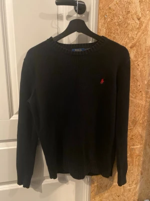 Svart stickad tröja från Polo Ralph Lauren - Klassisk svart stickad tröja från Polo Ralph Lauren med rund halsringning och den ikoniska röda logotypen broderad på bröstet. Tröjan har långa ärmar och ribbade muddar vid ärmslut och nederkant. Perfekt till jeans eller chinos.
