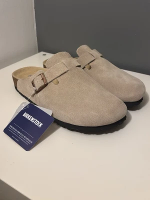 Ljus Beige Birkenstock tofflor i mocka - Snygga beige tofflor från Birkenstock i mjuk mocka med justerbart spänne och klassisk korksula. Perfekta för dig som vill ha både stil och komfort. Modellen har öppen häl och svart yttersula. Modellen är Boston. Storlek 37 😊