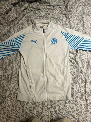 Vit Marseille träningsjacka Puma - Säljer en vit träningsjacka från Puma med Olympique de Marseille-logga på bröstet. Jackan har blåa ränder på ärmarna, hel dragkedja framtill och två fickor. Perfekt för dig som gillar fotboll och vill ha en sportig look.