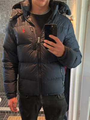 Mörkblå dunjacka från Polo Ralph Lauren - Säljer en mörkblå dunjacka från Polo Ralph Lauren med huva och dragkedja. Jackan har en röd broderad logga på bröstet och två sidofickor med dragkedja. Perfekt för kalla vinterdagar. Typ helt ny köptes i vintras och användes inte så mycket. Nypris 5500