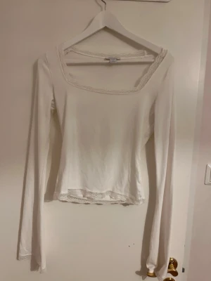 Vit långärmad topp med spetskant - Säljer en vit långärmad topp med fyrkantig ringning och fina spetsdetaljer vid halsen och nederkanten. Toppen har en slim passform och är perfekt att styla med jeans eller kjol.