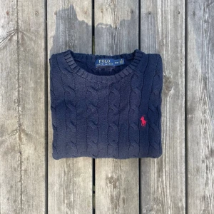 Ralph Lauren Cable Knit | Marinblå - StrI M - Kabelstickad mörkblå tröja från Polo Ralph Lauren med rund halsringning och den klassiska röda logotypen broderad på bröstet. Tröjan har långa ärmar och ribbade muddar vid ärmslut och nederkant. Perfekt för en stilren och avslappnad look. Skriv för bild hör gärna av dig vid minsta fundering eller om du bara har en dålig dag❤️ jag är alltid öppen till bud!