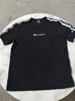 Svart Champion t-shirt med logga - Svart t-shirt från Champion med vit logga på bröstet och snygga band med Champion-tryck längs axlarna. Klassisk rund hals och korta ärmar. Tillverkad i mjuk bomull för skön känsla hela dagen.
