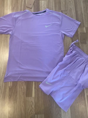 Lila träningsset från Nike - Säljer ett snyggt träningsset från Nike i ljuslila färg. Setet består av en kortärmad t-shirt och matchande shorts, båda med Nike-logga i vitt. Perfekt för gymmet eller löprundan. Materialet är lätt och andas bra.