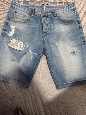 Ljusblå slitna jeansshorts - Säljer ett par ljusblå jeansshorts med slitningar och råa kanter. Shortsen har fem fickor, dragkedjegylf och coola detaljer som trasiga partier och patch på baksidan. Perfekta för en avslappnad och trendig look.