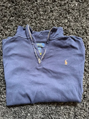 Blå långärmad tröja från Polo Ralph Lauren - Mörkblå långärmad tröja från Polo Ralph Lauren med halv dragkedja och klassisk broderad logga på bröstet. Tröjan har hög krage och är tillverkad i mjukt bomullsmaterial. Perfekt för en avslappnad och stilren look.