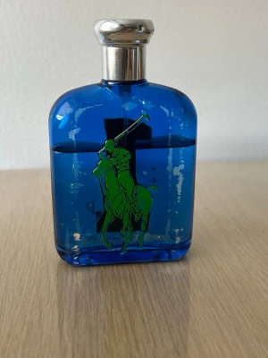 Blå parfymflaska med gul etta - En cool parfymflaska i blått glas med silverfärgat lock. På framsidan syns en grön polospelare och på baksidan en stor gul siffra 1. Flaskan har en sportig och ungdomlig design som sticker ut i badrumshyllan.