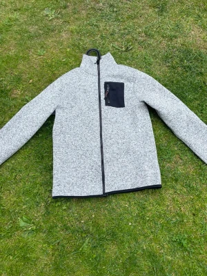 Ljusgrå cardiganjacka med dragkedja - Snygg ljusgrå cardiganjacka med hel dragkedja framtill och svart bröstficka med dragkedja. Jackan har hög krage och är tillverkad i ett mjukt, stickat material. Perfekt för lager på lager och chillare dagar.
