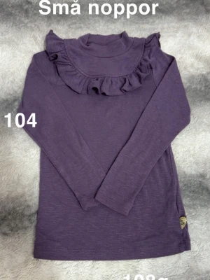 Lila långärmad topp med volang - Lila långärmad topp med hög krage och volangdetalj över bröstet. passar perfekt till vardagslooken. Enkel men ändå söt design som ger en trendig touch.🌸 Små noppor. Storlek 104