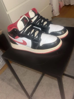 Air Jordan 1 Mid svart/röd/vit - Säljer ett par Air Jordan 1 Mid sneakers i svart, rött och vitt. Skorna har klassisk Jordan-logga på plösen, svart läderdetaljer, vit tåbox och sidor samt röd swoosh och häl. Yttersulan är röd och mellansulan vit. Perfekta för dig som gillar streetwear och ikoniska sneakers.