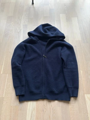 Mörkblå stickad casmre hoodie - Mysig mörkblå casmre hoodie i stickat material med dragkedja framtill och huva. Tröjan har långa ärmar och ribbade muddar vid ärmslut och nederkant. Perfekt för en avslappnad och stilren look. 