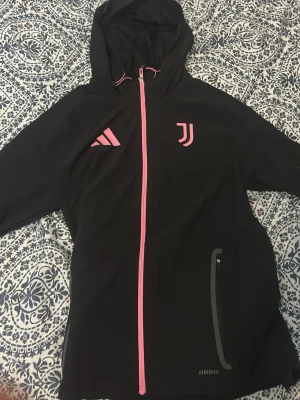 Svart Juventus hoodie med rosa detaljer - Snygg svart hoodie från Adidas med Juventus-logga och rosa detaljer. Dragkedja och loggor i rosa, huva och ficka med dragkedja. Perfekt för träning eller chill. Materialet är troligtvis polyester med Aeroready-teknologi helt ny fast var för liten för mig 