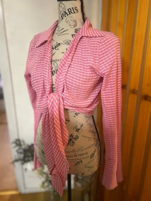 Rosa randig knytblus med krage - Trendig rosa blus med vita ränder och lång ärm. Blusen har en snygg krage och knyts framtill för en cool och avslappnad look. Perfekt att styla med jeans eller kjol för en fräsch outfit.