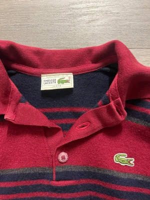 Randig röd och marinblå Lacoste skjorta - Säljer en klassisk randig skjorta från Lacoste i rött och marinblått. Skjortan har krage, knappar framtill och den ikoniska gröna krokodilloggan på bröstet. 