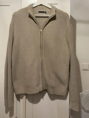 Beige stickad ziptröja från 157 - Säljer en beige stickad tröja från 157 med dragkedja framtill och hög krage. Tröjan har lång ärm och ribbade muddar vid ärmslut och nederkant. Perfekt för lager på lager och enkel att matcha med olika outfits.