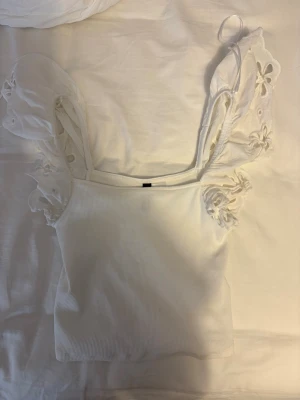 Söt volang topp från hm - Snygg vit ribbad topp med breda axelband och volangärmar i broderad mesh. Toppen har fyrkantig ringning och är figurnära i modellen. Perfekt för en trendig och fräsch look.