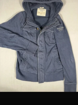 Blå cardigan från Abercrombie & Fitch - Snygg blå hoodie från Abercrombie & Fitch med dragkedja, ribbade muddar och fyra fickor framtill. Märkeslogga på bröstet och huva med snörning. Perfekt för en avslappnad och cool stil. Tillverkad i mjukt bomullsmaterial. Storlek L men sitter som M