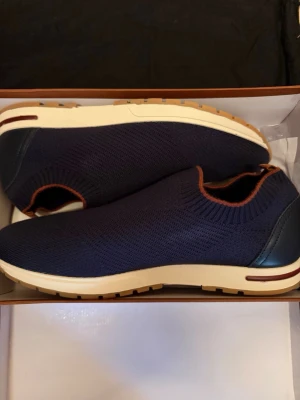 Loro Piana Marinblåa - Snygga marinblå slip-on sneakers från Loro Piana med stickad ovandel och kontrasterande bruna detaljer. Skorna har vit sula med bruna inslag och mörkblå hälparti i läder. Perfekta för en avslappnad och trendig look.