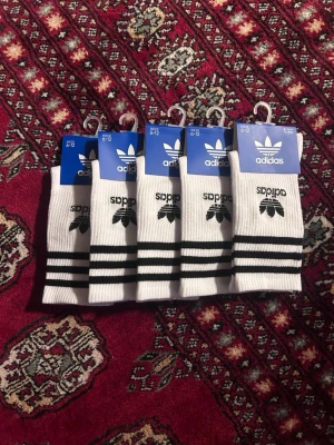Vita Adidas strumpor med svarta ränder 5st  - Klassiska vita Adidas strumpor med svarta ränder och Adidas logga på sidan. Ribbad kant upptill för skön passform. Perfekta till sneakers och streetwear. Säljs i flerpack.