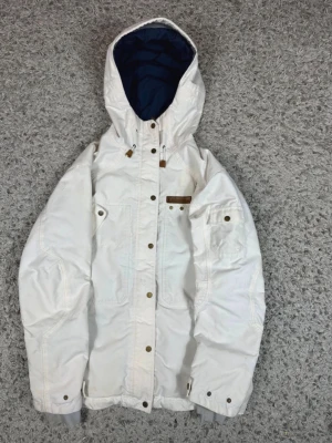 Vit parkas med huva och detaljer - Snygg vit parkas med justerbar huva och mörkblått innerfoder. Jackan har flera fickor med tryckknappar, dragsko i huvan och detaljer i brunt läder. Perfekt för kalla dagar och har en klassisk, stilren look.