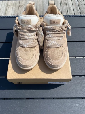 sneakers UGG Lowmel - Snygga beige sneakers från UGG med grov sula och detaljer i mocka och mesh. Insidan är fodrad med mjuk fuskpäls för extra komfort. Skorna har matchande beige skosnören och kommer med ett extra par. Perfekta för dig som vill ha både stil och komfort.