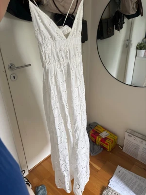 Vit långklänning med broderade detaljer - Vit långklänning med smala axelband och v-ringning. Klänningen har broderade hålmönster och en smockad midja för en bekväm passform. Perfekt för varma dagar och somriga tillfällen.