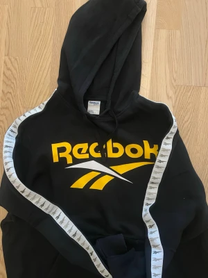 Svart Reebok hoodie med gula detaljer - Snygg svart hoodie från Reebok med stor gul logga och vita detaljer på ärmarna. Huva med dragsko och magficka framtill. Ärmarna har vita band med grått mönster längs sidorna. Perfekt för en sportig och avslappnad stil.