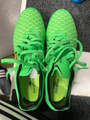 Nike Magista gröna fotbollsskor - Säljer ett par Nike Magista fotbollsskor i neon-grönt med svart och orange swoosh på sidan. Skorna har snörning, mönstrad ovandel och fasta dobbar i plast och metall för bra grepp på planen. Insidan är svart och grön. Perfekta för dig som vill sticka ut på fotbollsplanen!