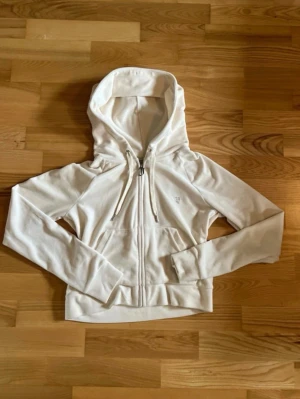 Vit Juicy Couture hoodie med dragkedja - Säljer en vit hoodie från Juicy Couture med dragkedja framtill, fickor och huva med snören. Baksidan har ett stort hjärtmotiv med krona och texten 'Juicy Couture' i glittriga stenar. Storlek m men upplevs väldigt liten❤️