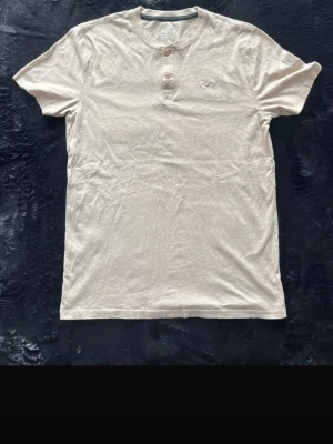 Hollister henley  - Snygg beige t-shirt från Hollister med två knappar vid halsen och rundad krage. T-shirten har korta ärmar och en liten broderad logga på bröstet. Tillverkad i mjuk bomull för skön känsla och enkel stil. Den är något mörkare i annat ljus 